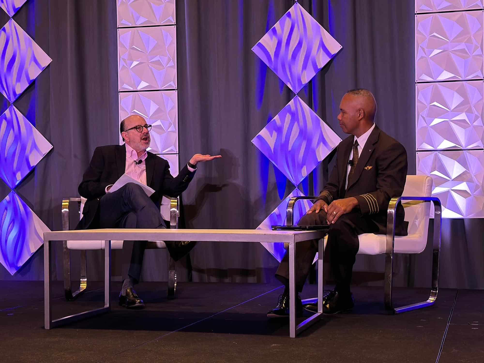 Two men sit on stage in chairs, talking during an event. One man gestures while speaking, and the other, wearing a dark suit with pilot insignia, listens. The background features geometric panels lit with purple light.