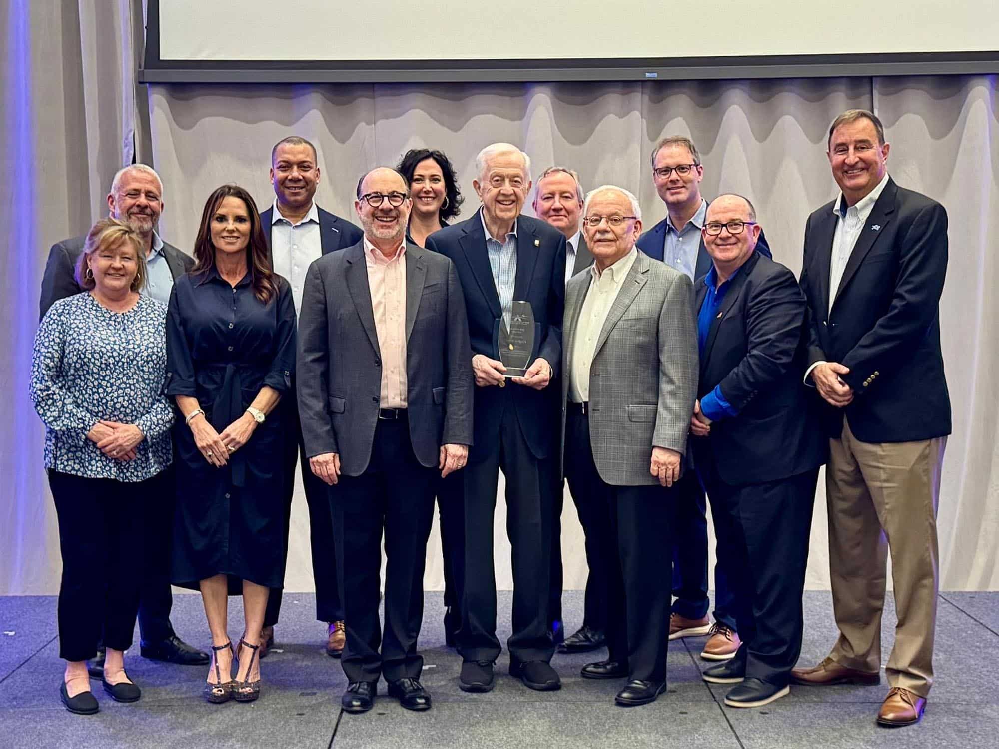 A group of twelve adults, dressed in business attire, stand together on a stage. The older man in the center holds an award and everyone is smiling toward the camera in a formal, celebratory setting.