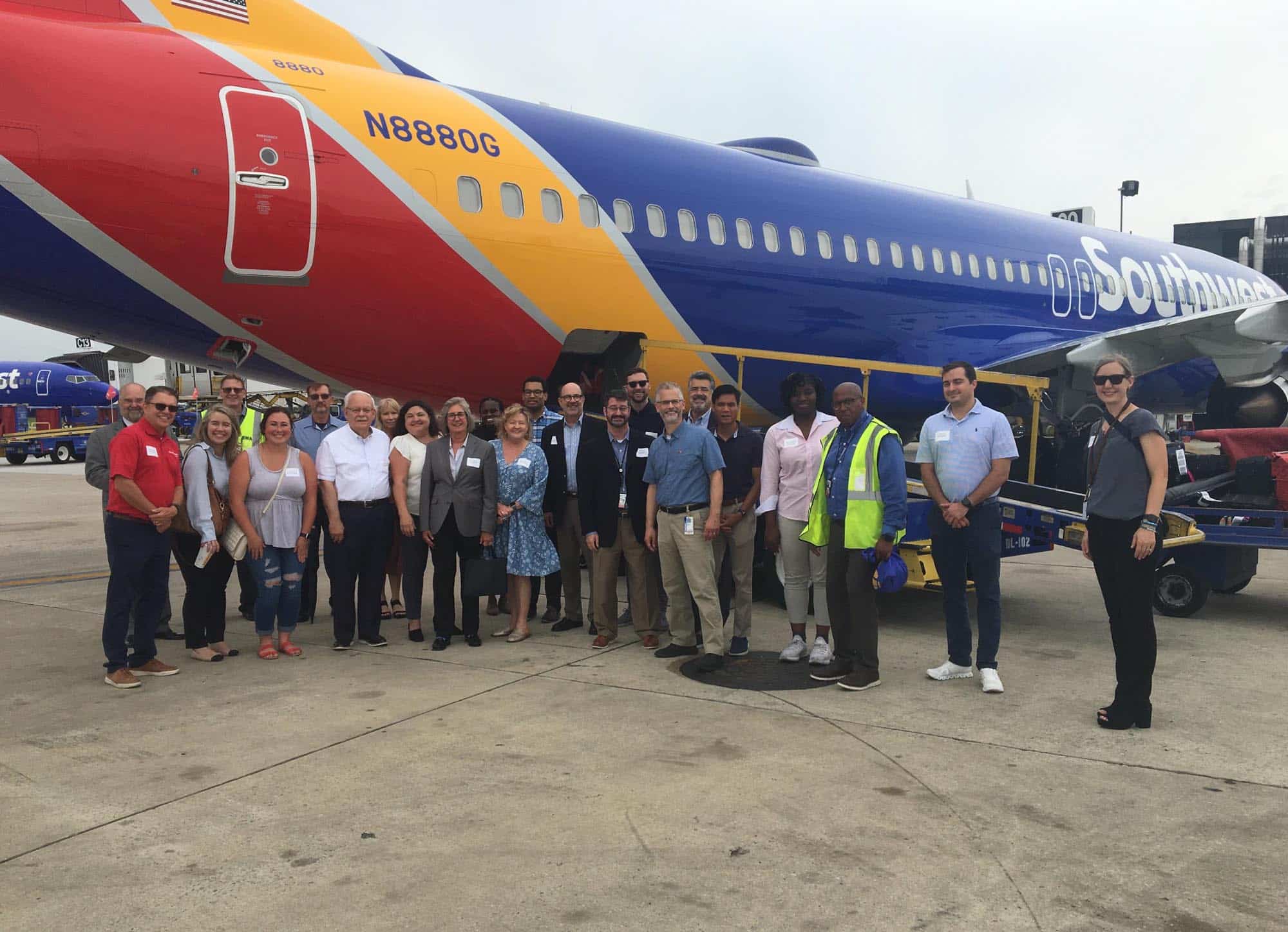 A group of people pose and smile together in front of a Southwest Airlines plane on an airport tarmac, with some wearing business attire and others in safety vests.