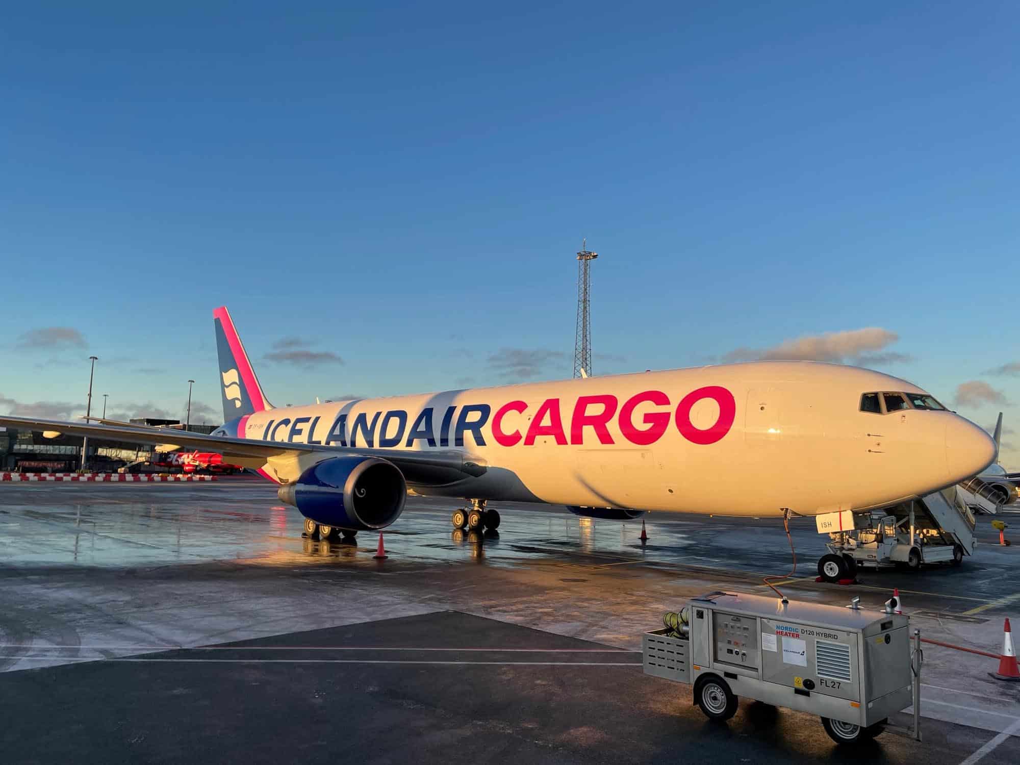 An Icelandair Cargo airplane is parked on the tarmac near an airport terminal at sunrise or sunset, with ground service equipment and orange cones nearby. The sky is clear with a few clouds.