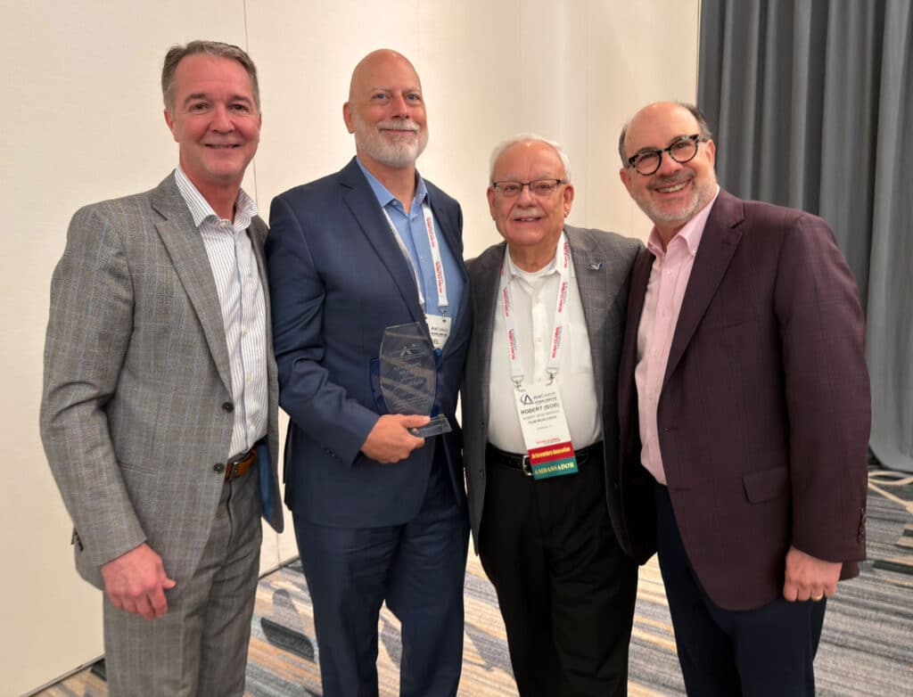 noel Four men in business attire stand smiling together indoors; one is holding a Jim Foster Award. They each have conference name badges and are standing on a patterned carpet with a white and gray backdrop.