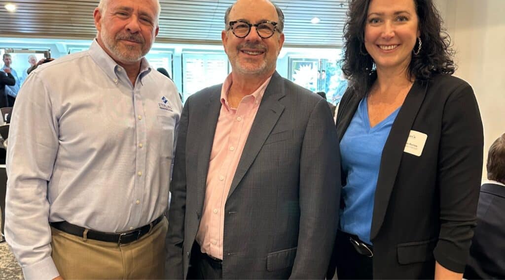 Three adults stand indoors, smiling at the camera. The two men wear business attire: one in a blue dress shirt and khakis, the other in a pink shirt and suit. A woman in a blazer and blue top stands beside them. Other people are visible in the background.