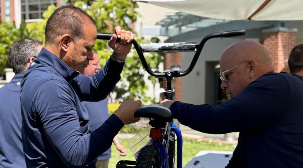 Two men in navy blue shirts assemble a bicycle outdoors on a sunny day, focusing on the handlebars and seat, with greenery and a building visible in the background.