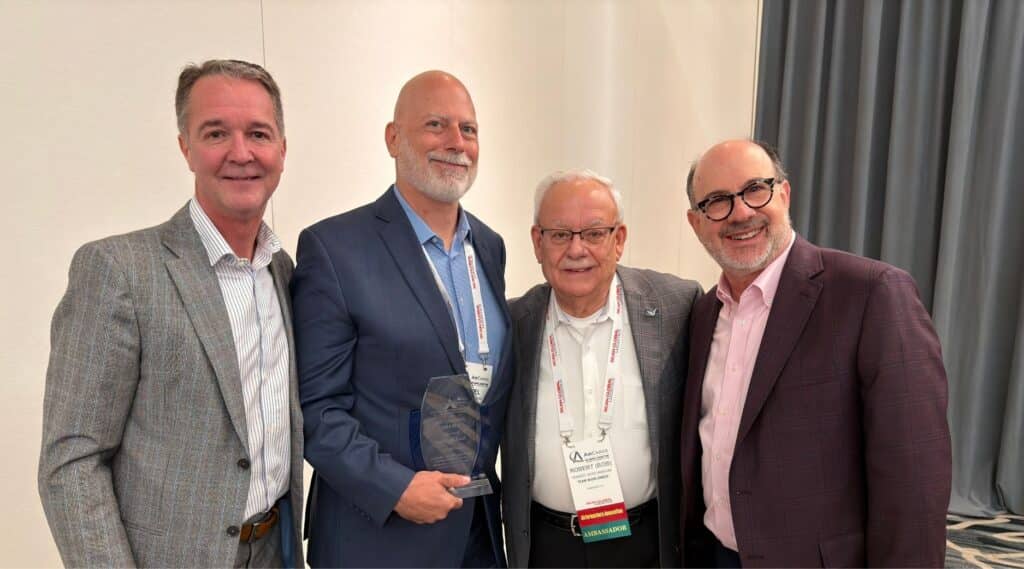 Four men in business attire stand smiling for a photo; one is holding a glass award. They are indoors, in front of a white and gray background, wearing conference name badges.