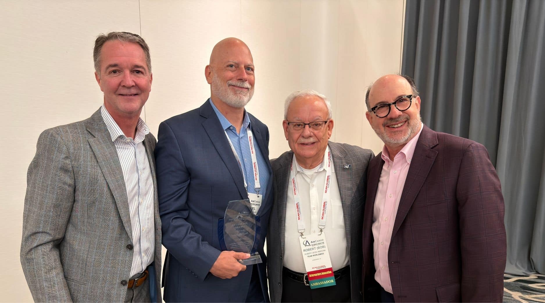 Four men in business attire stand smiling for a photo; one is holding a glass award. They are indoors, in front of a white and gray background, wearing conference name badges.