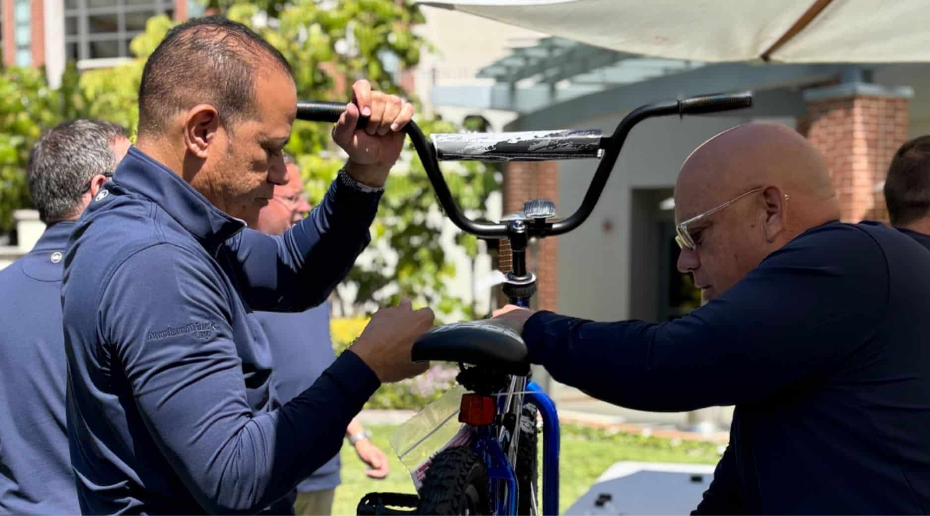 Two men in navy blue shirts assemble a bicycle outdoors on a sunny day, focusing on the handlebars and seat, with greenery and a building visible in the background.